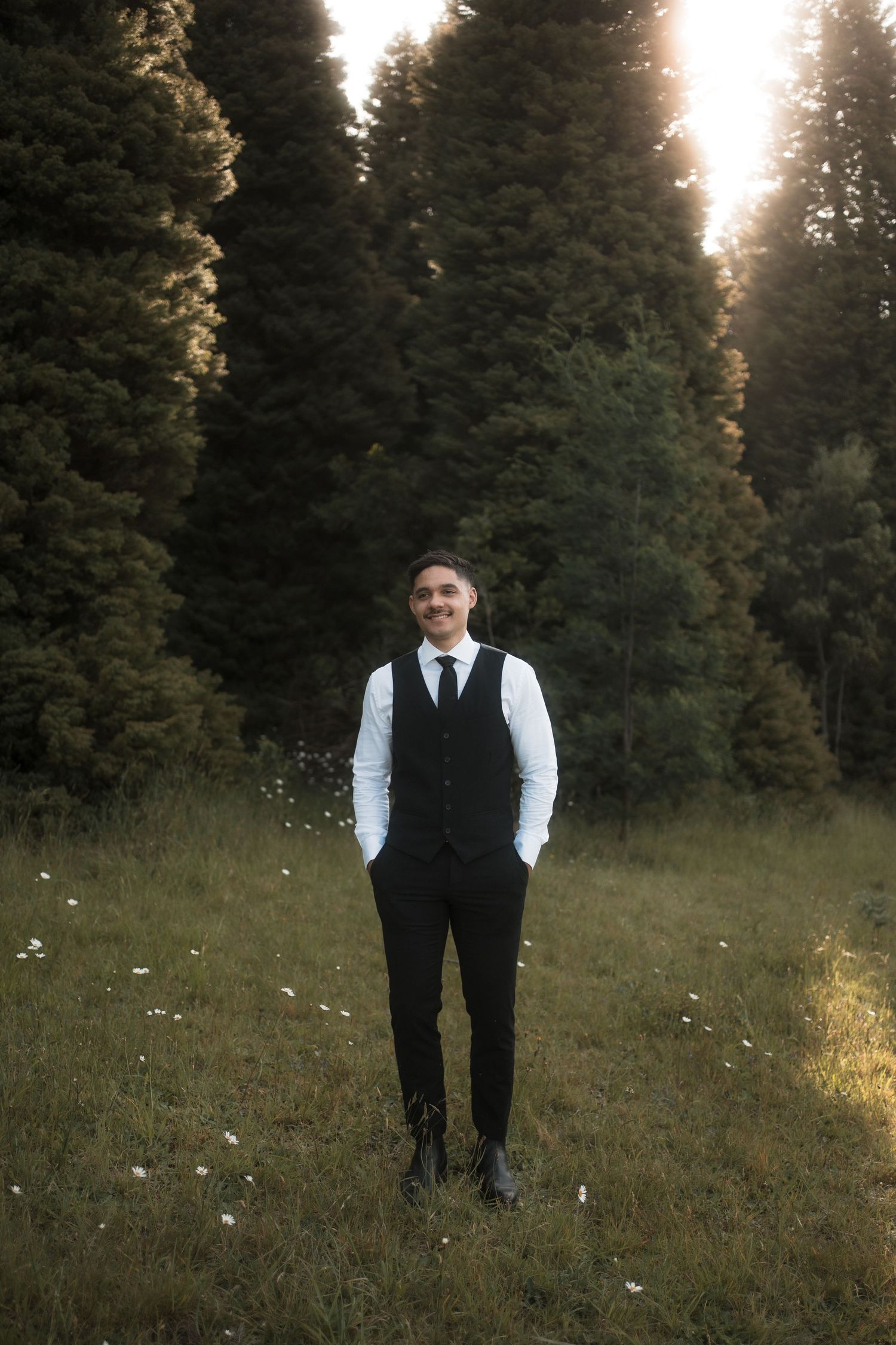 A person in formal attire stands in a misty meadow at sunset among evergreen trees.