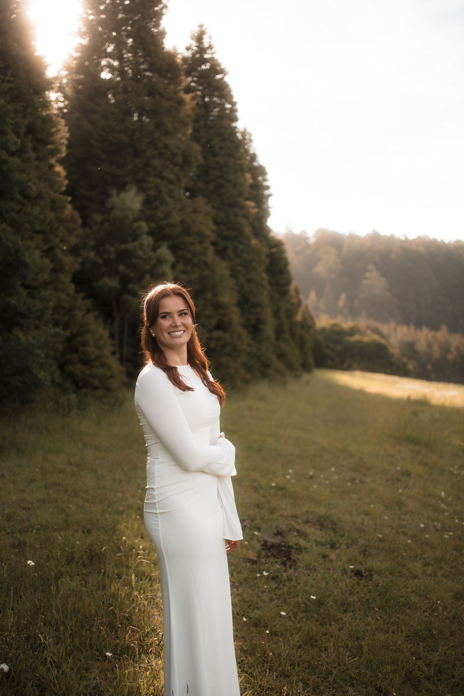A bride in a sleek white long-sleeve gown poses in a scenic mountain meadow at sunset.