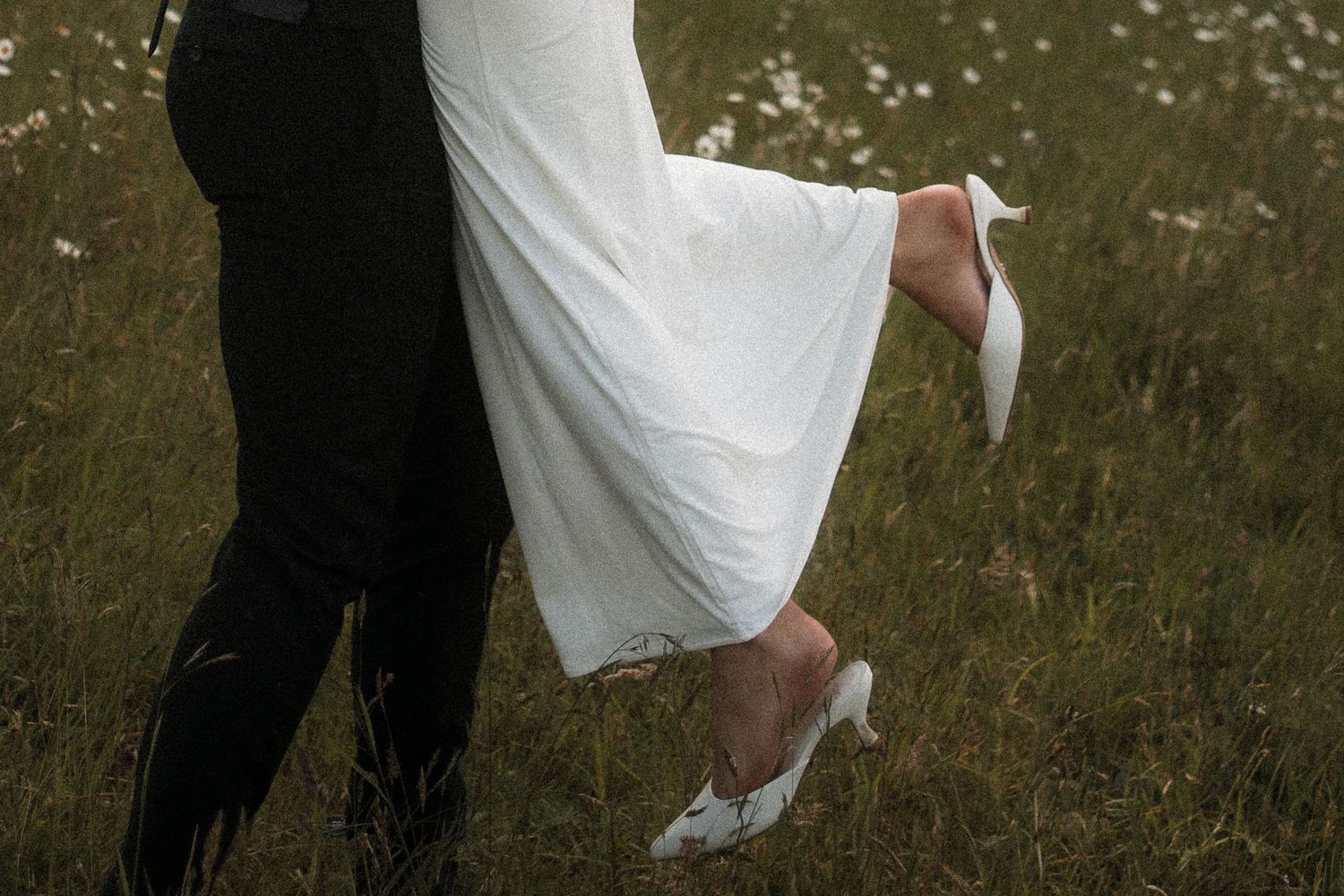 Close-up of bride and groom's legs in a grassy field, with white wedding dress and black suit visible.