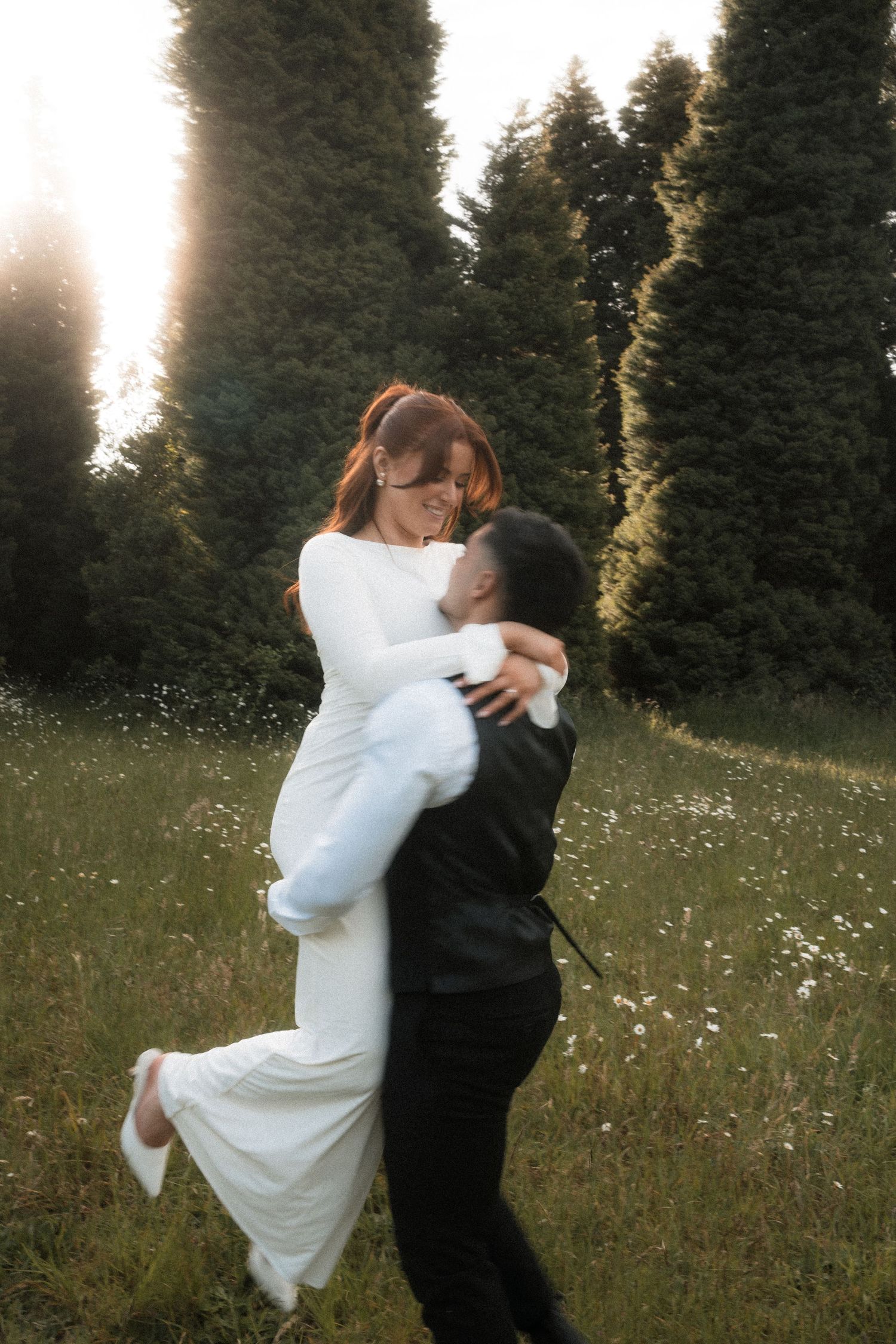 A couple embraces in a sunlit meadow with wildflowers, the bride wearing a flowing white dress against evergreen trees.