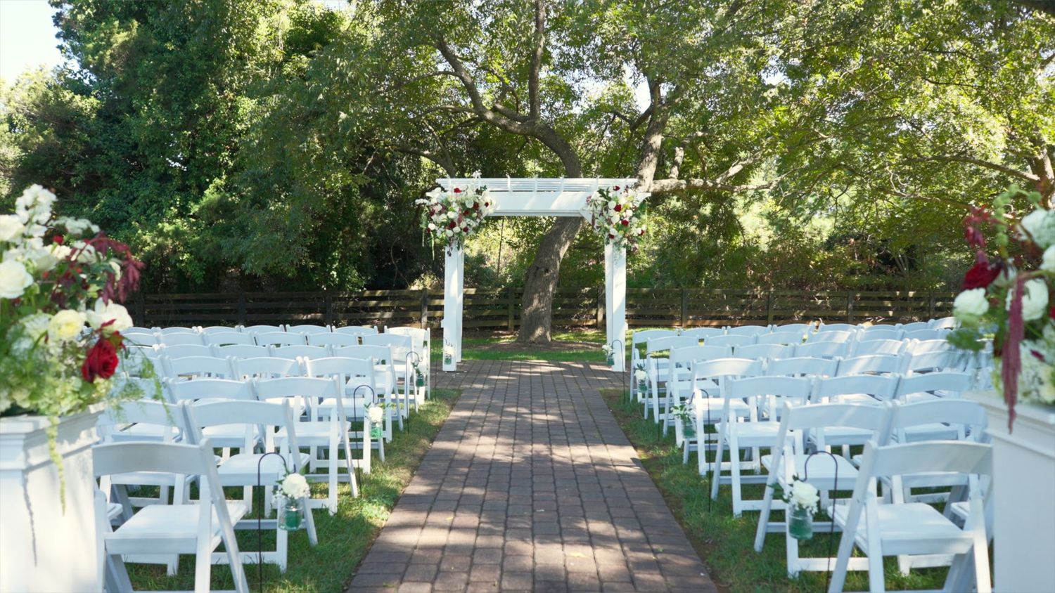 White folding chairs line a brick pathway leading to a garden wedding ceremony setup under trees.