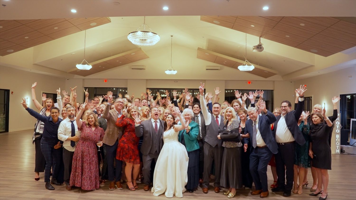 Large group of wedding guests and newlyweds pose together with raised arms celebrating in an elegant reception venue.