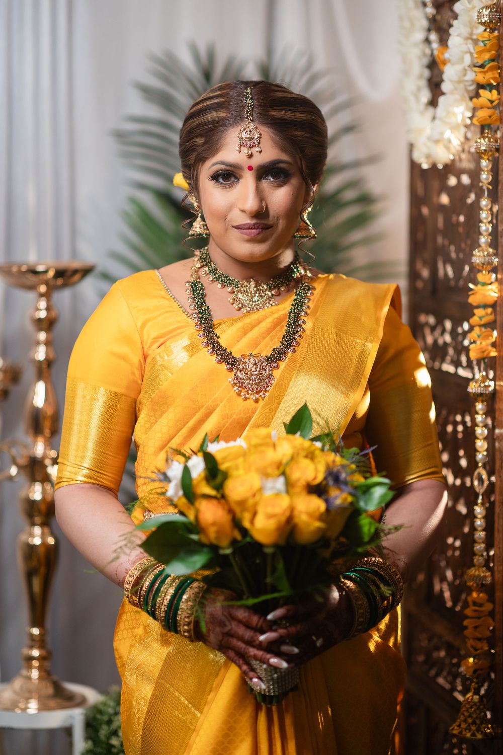 Indian bride in yellow silk saree holds yellow roses while adorned with traditional jewelry and decorative elements.