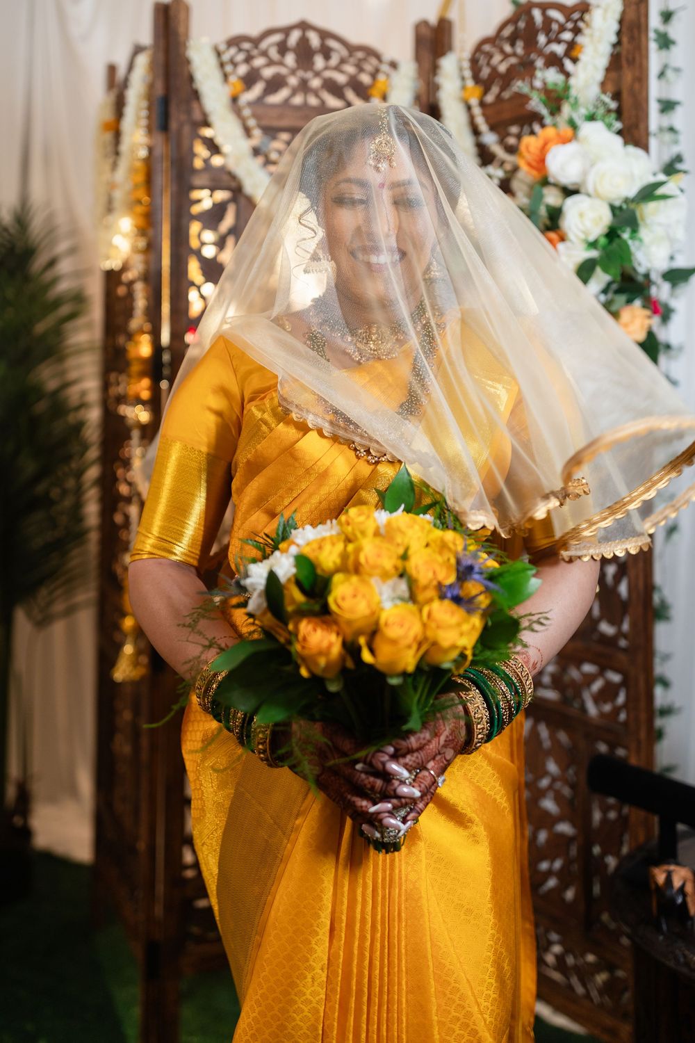 Traditional bride in vibrant yellow dress holds yellow and white bouquet with delicate veil and ornate backdrop.