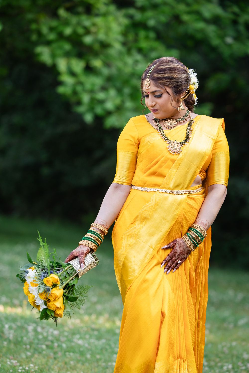 Bride in marigold yellow sari poses gracefully in outdoor garden setting with natural greenery.