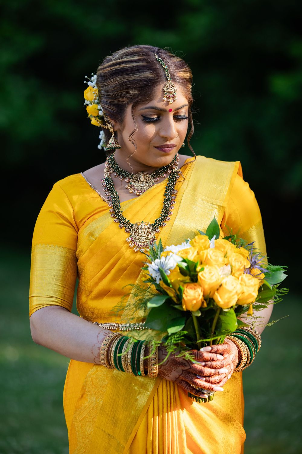 Bride in vibrant yellow dress holding yellow and white floral bouquet against green garden backdrop.