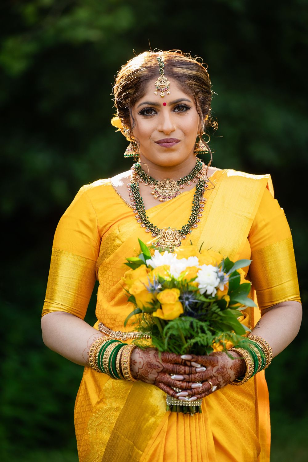 Person in yellow silk saree holding yellow and white flowers with mehndi-adorned hands.