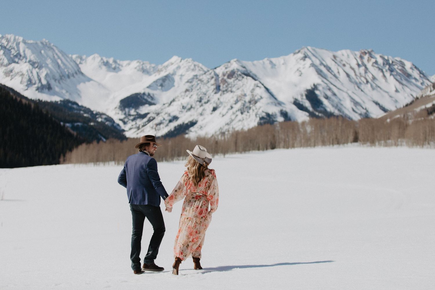 Couple holding hands in a mountain meadow with red wildflowers and snow-capped peaks in the background.