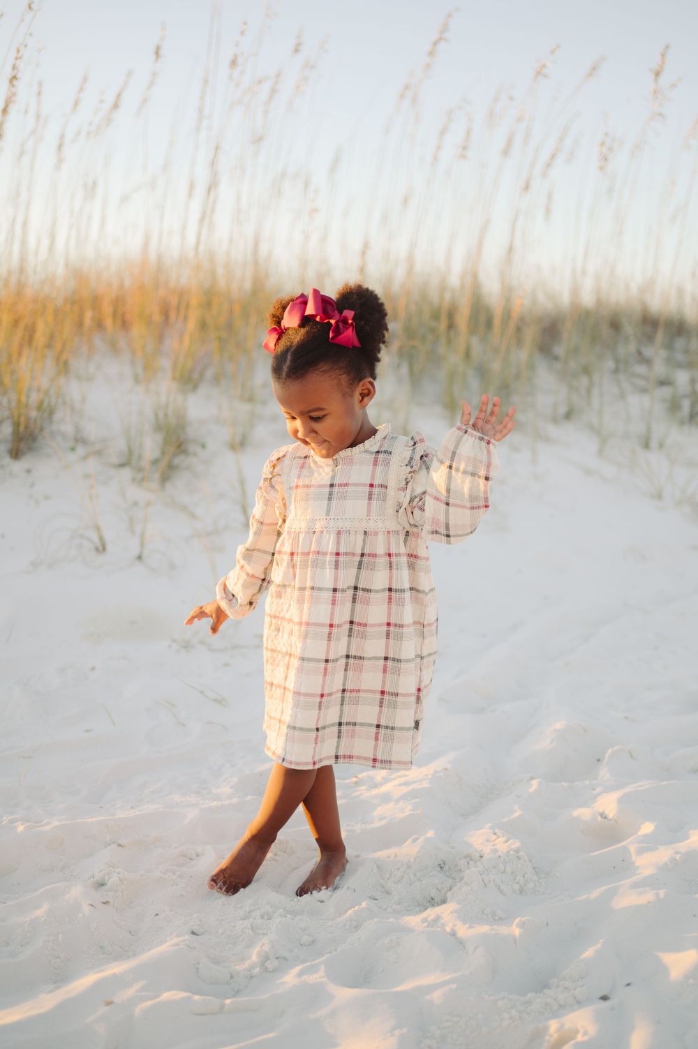 A small child wearing a plaid dress and red bows plays joyfully in white sand dunes at sunset.