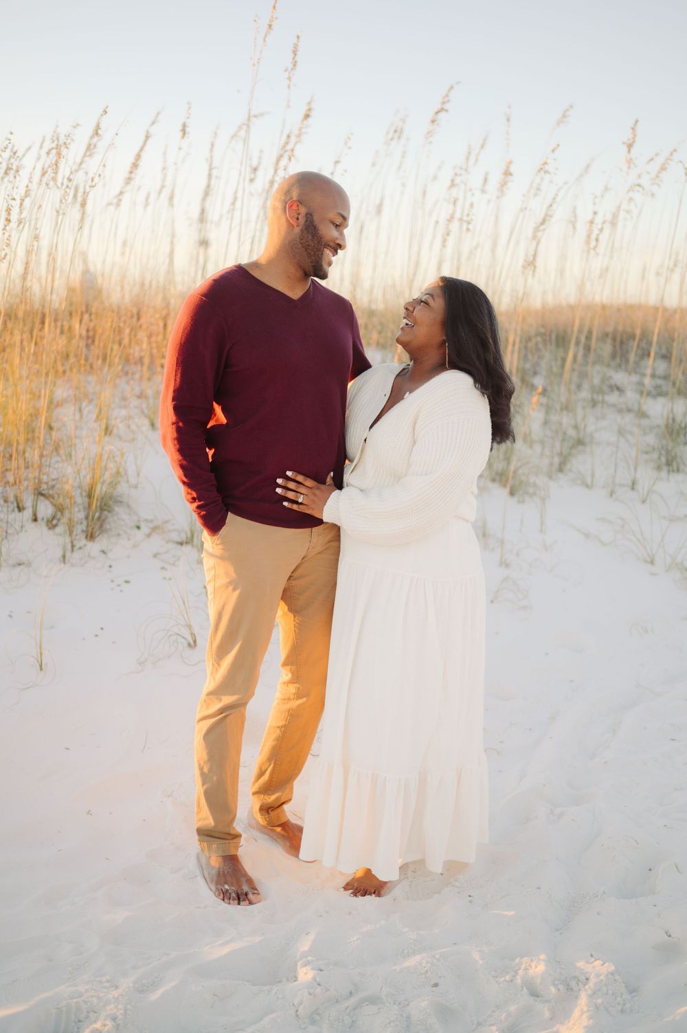 A couple embraces on a white sandy beach with sea oats swaying in the golden sunlight.