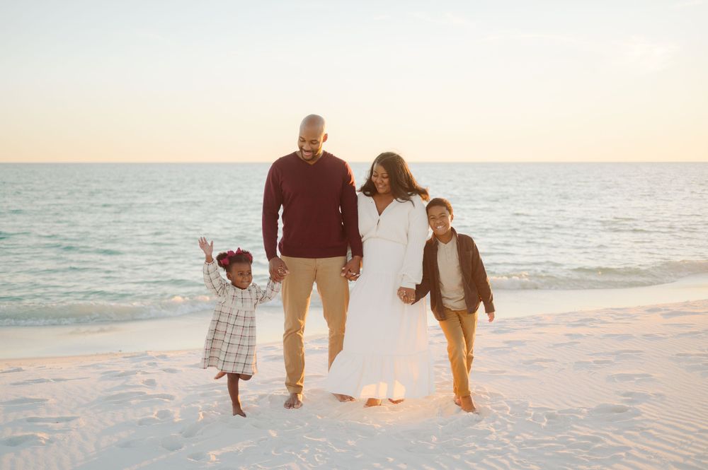 A family of three walks together on a white sandy beach at sunset with the ocean in the background.