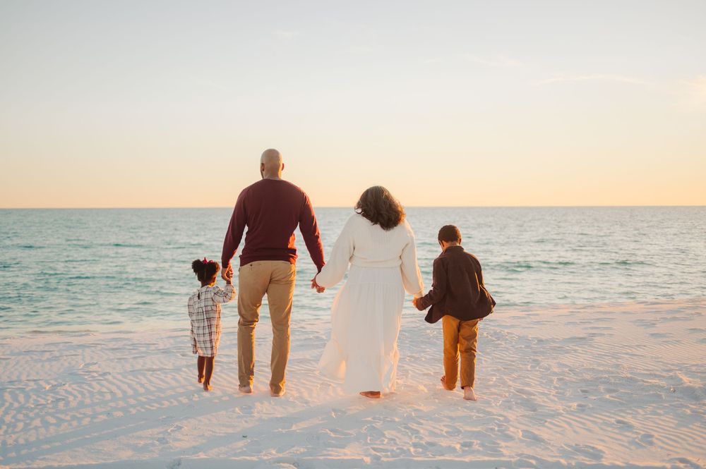 Family walking together on white sandy beach at sunset while holding hands and looking at ocean.