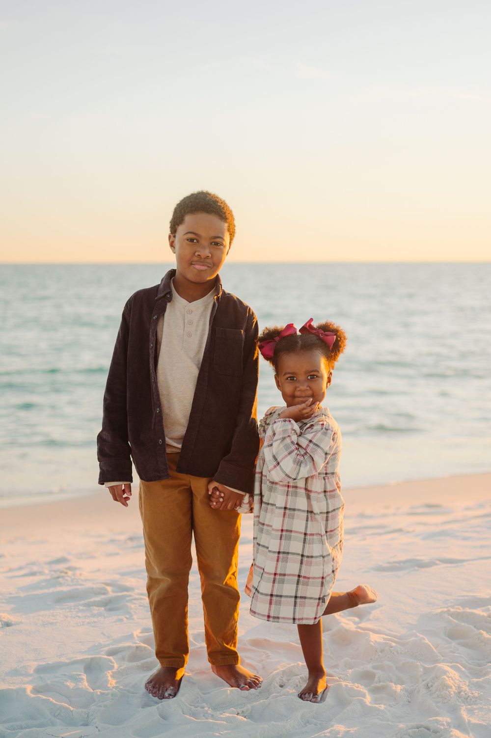 Two people stand barefoot on white sand beach at sunset with ocean waves in the background.