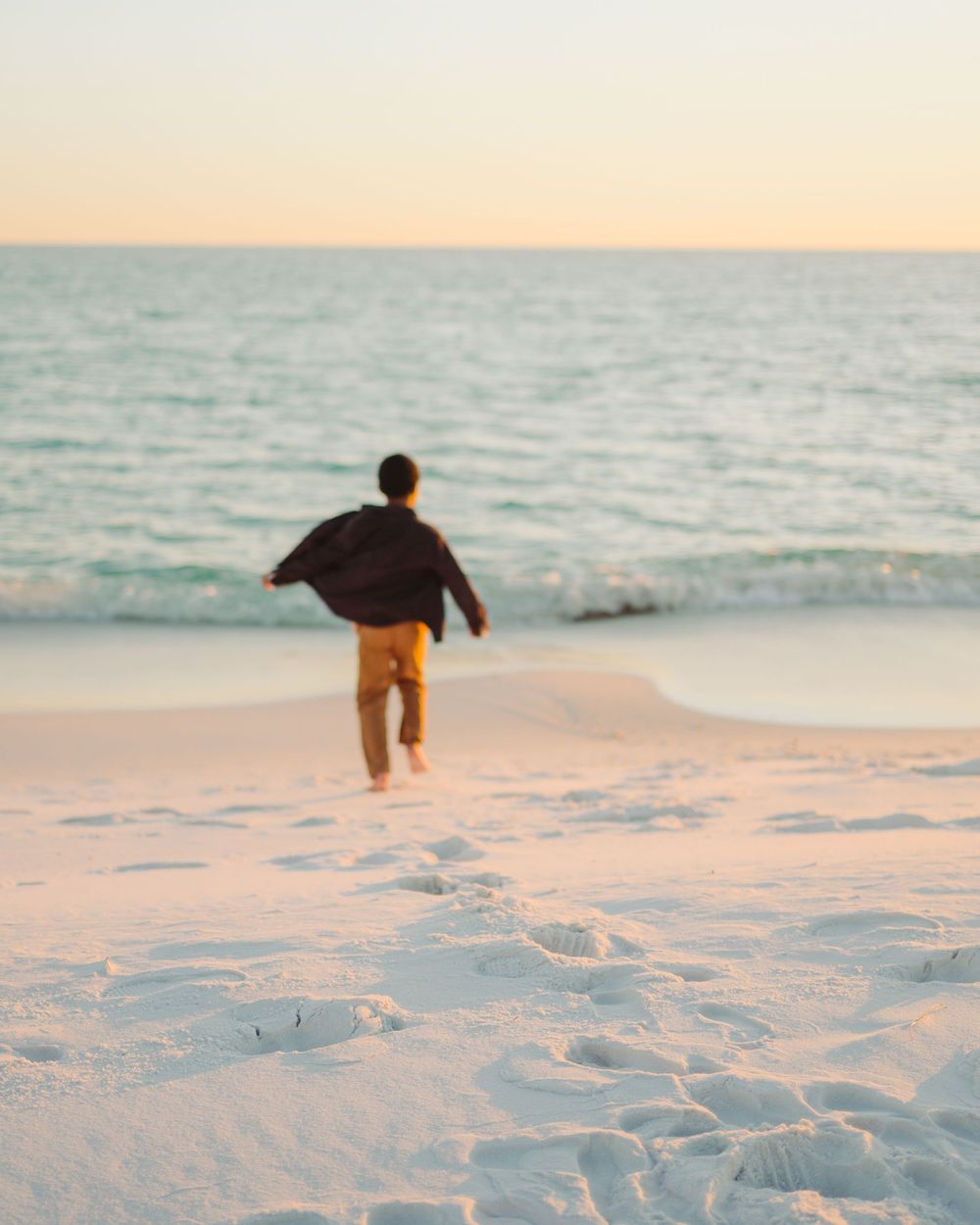 A person in dark clothing runs joyfully across pristine white sand beach at sunset with turquoise ocean waters behind.