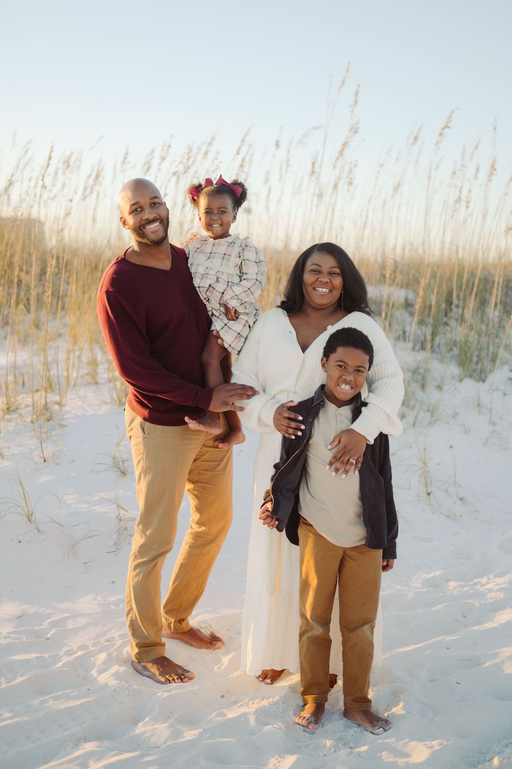 A family poses together barefoot on a white sand beach with sea oats swaying in the background during sunset.
