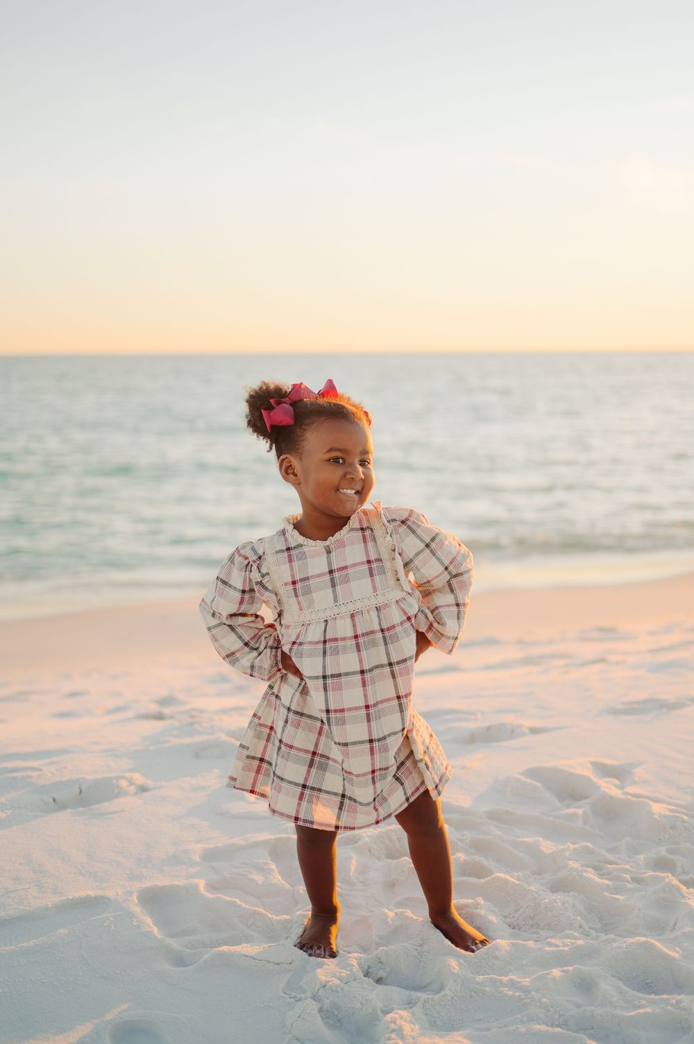 A toddler in a plaid dress dances joyfully on a pristine white sand beach at sunset.