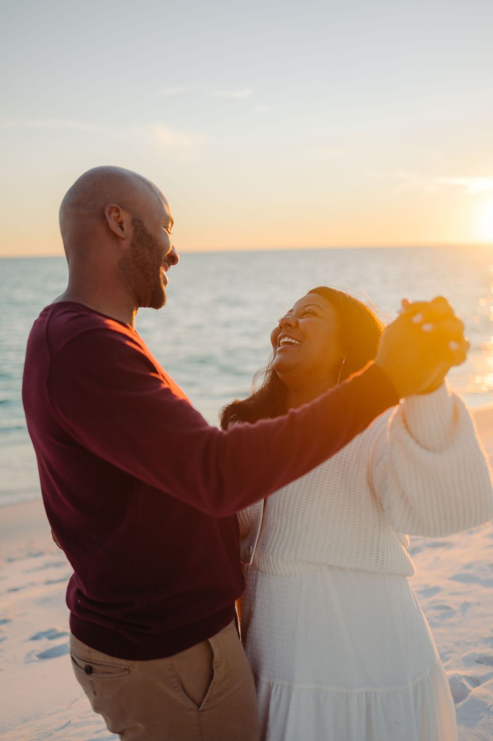 A couple embraces on a beach at sunset with golden sunlight streaming behind them.
