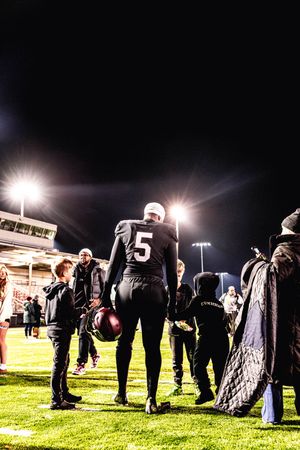 Spectators gather on a dark field at night with bright stadium lights casting dramatic shadows on the grass.