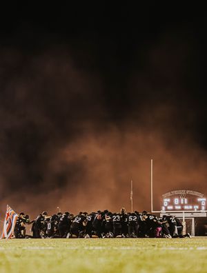 A nighttime football game showing a line of players and coaches on the sideline with stadium lights illuminating the field.