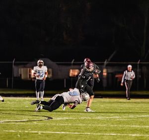 Football player evades a diving tackle attempt during a nighttime game as officials and players look on from the sideline.