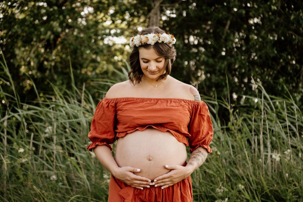 Pregnant woman in off-shoulder rust dress with floral crown looking down at bare baby bump in tall grass field.