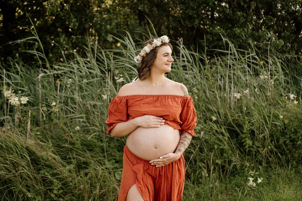 Pregnant woman wearing floral crown and rust orange off-shoulder dress cradling bump while standing in green meadow.