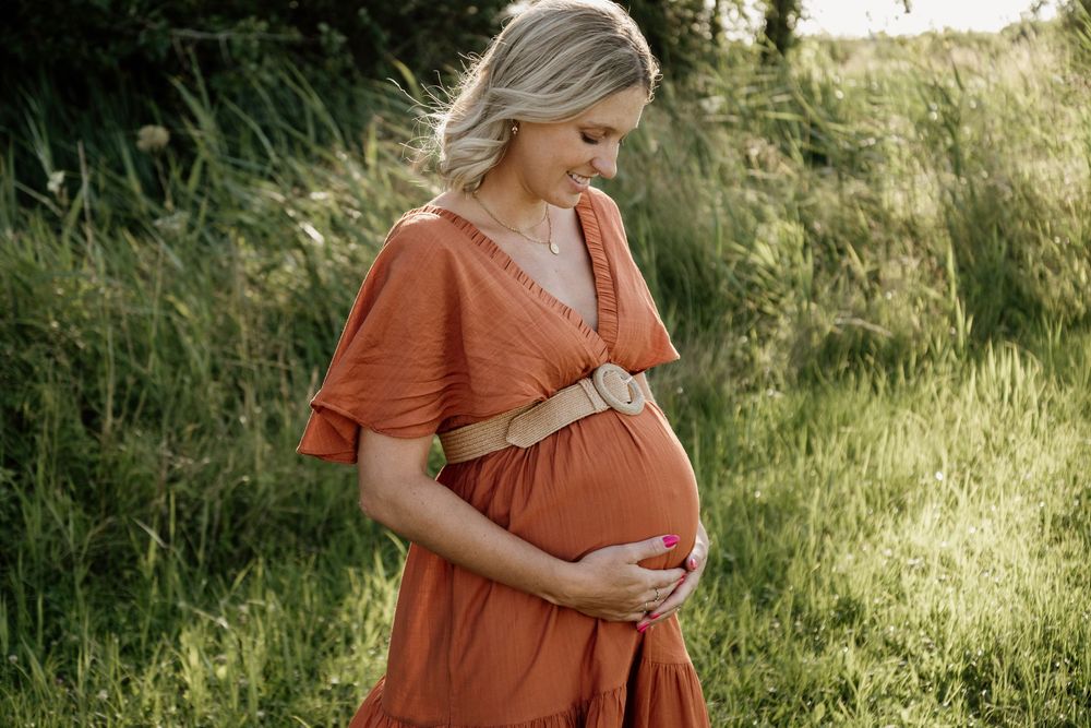 Pregnant woman in rust orange dress with belt gently holding baby bump while standing in green grassy field.