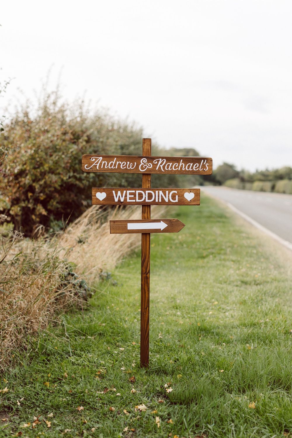 Three wooden directional wedding signs with arrows point the way along a grassy roadside path.