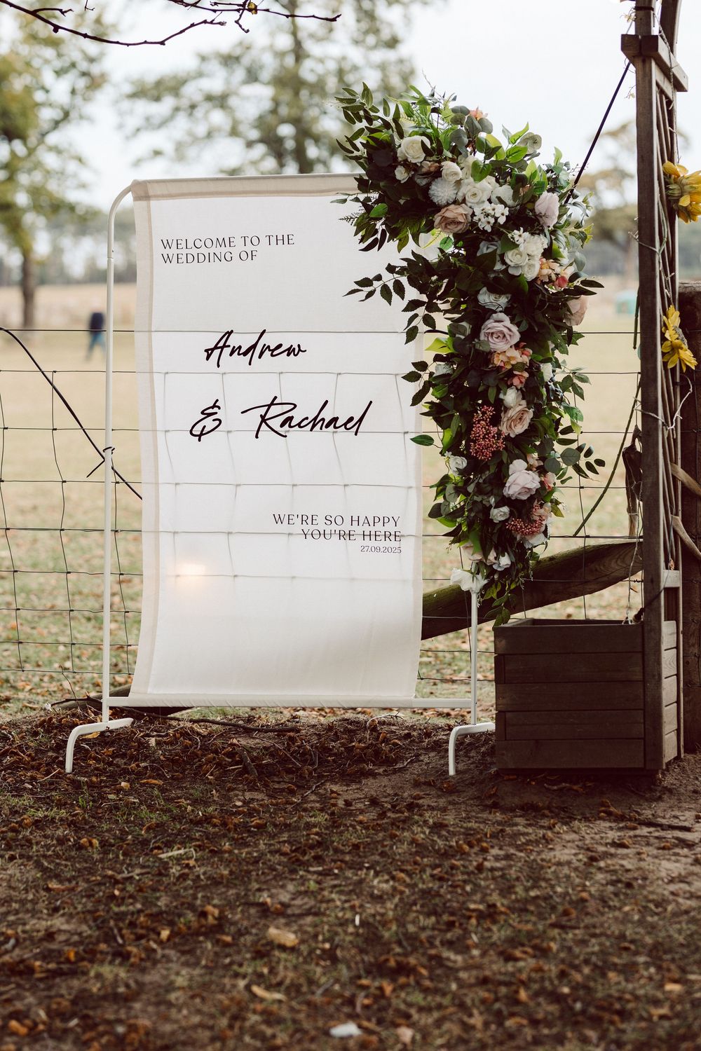 Rustic wooden welcome signs with elegant script lettering displayed on brown soil with scattered leaves and floral decor.