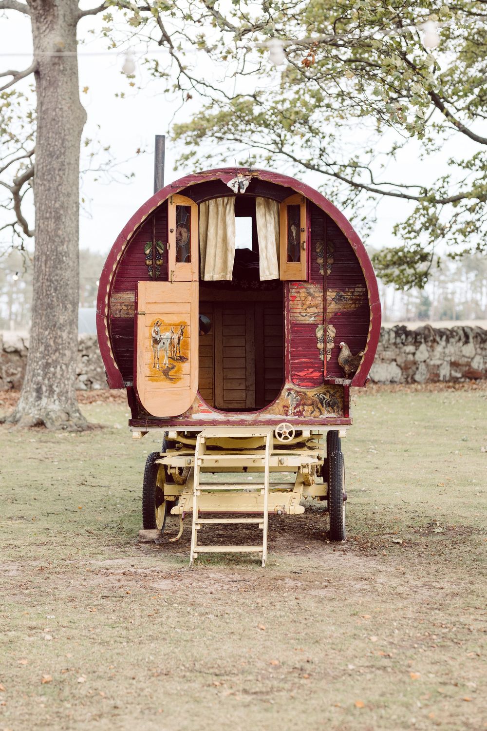 Three cozy burgundy-colored gypsy wagon campers with wooden steps and rustic details sit in a peaceful woodland setting.