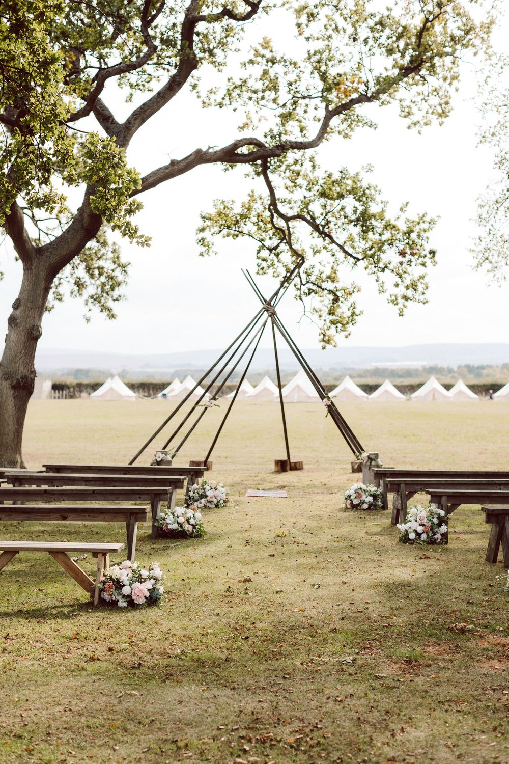 A rustic outdoor wedding setup with wooden benches and floral arrangements under a large tree in a scenic field.