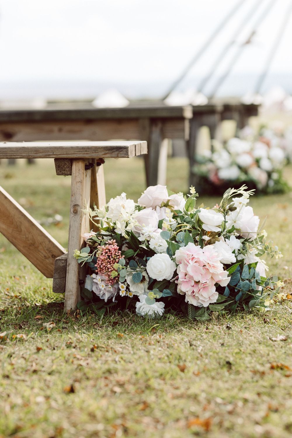 A rustic wooden bench decorated with lush pink and white floral arrangements and greenery for an outdoor wedding ceremony.