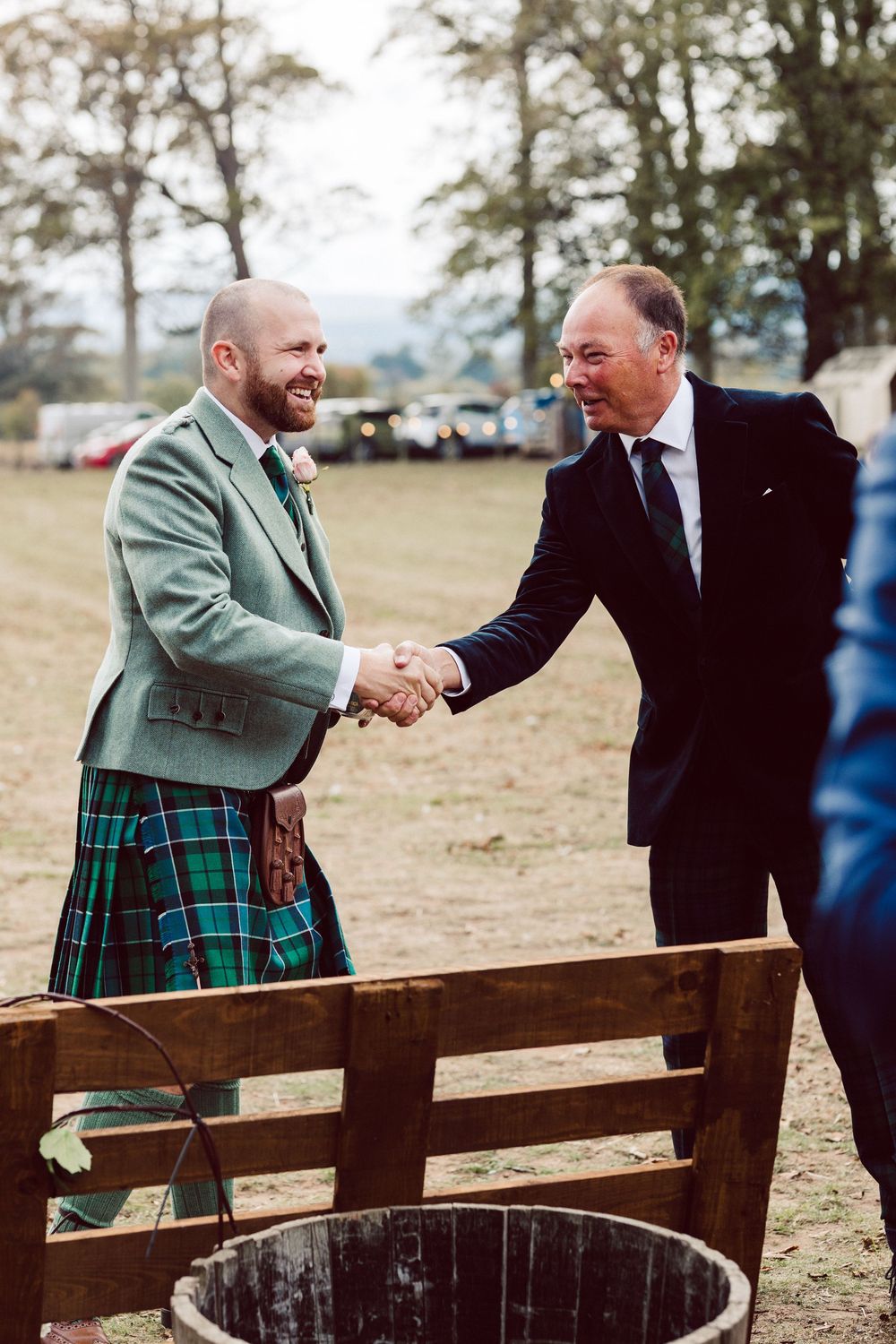 Two men in formal attire, one wearing a green jacket and kilt, shake hands and smile while seated on wooden benches outdoors.