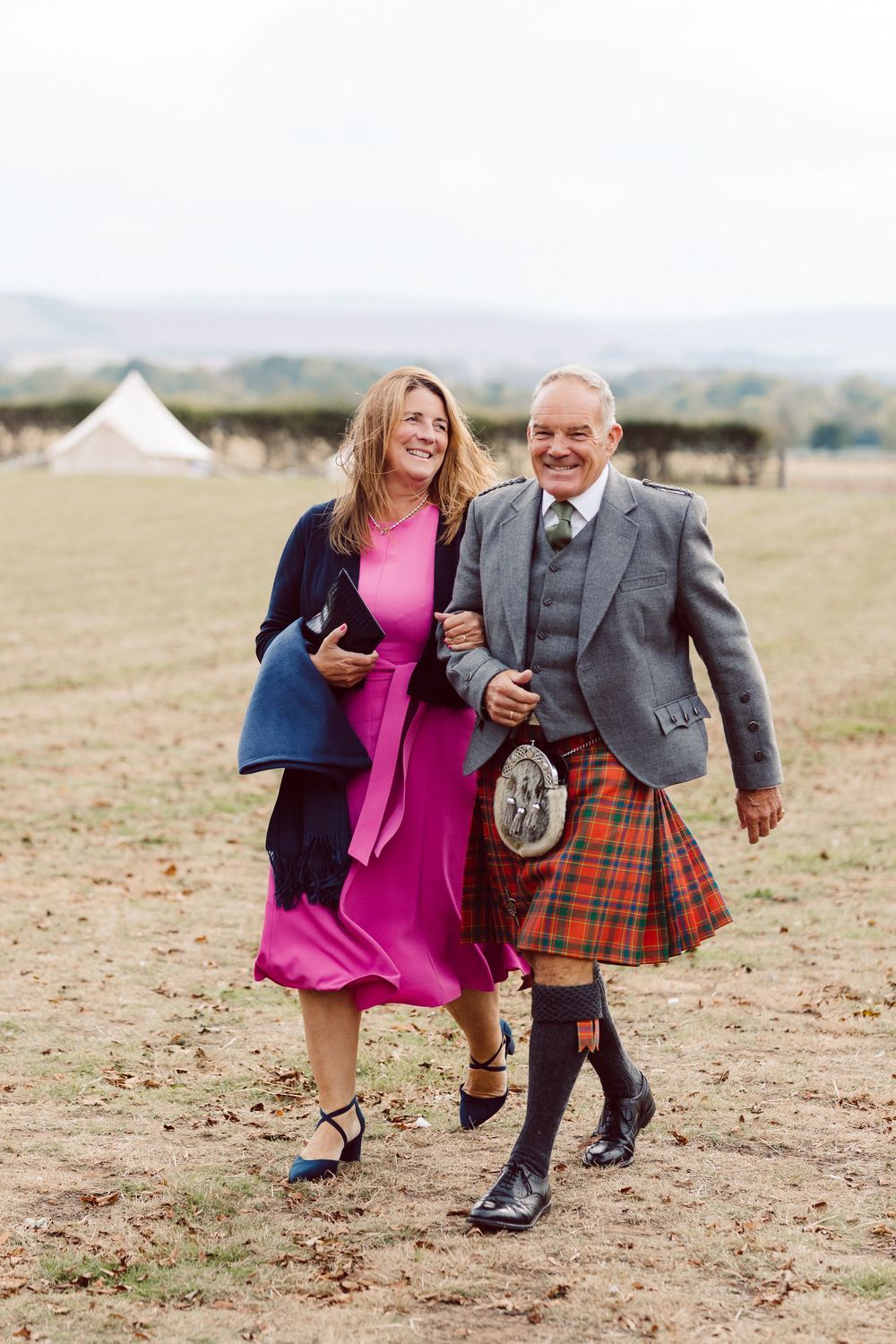A couple in Scottish attire dances joyfully together on a grassy field during a celebration.
