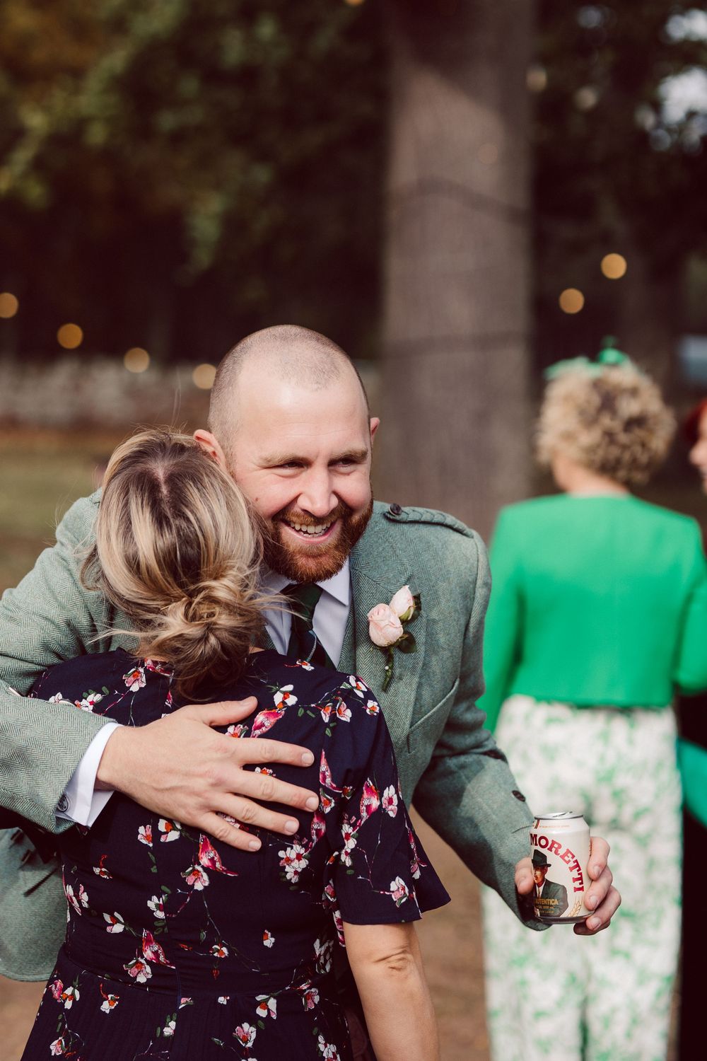 A couple shares an emotional embrace at an outdoor wedding reception with green bridesmaids dresses visible in background.