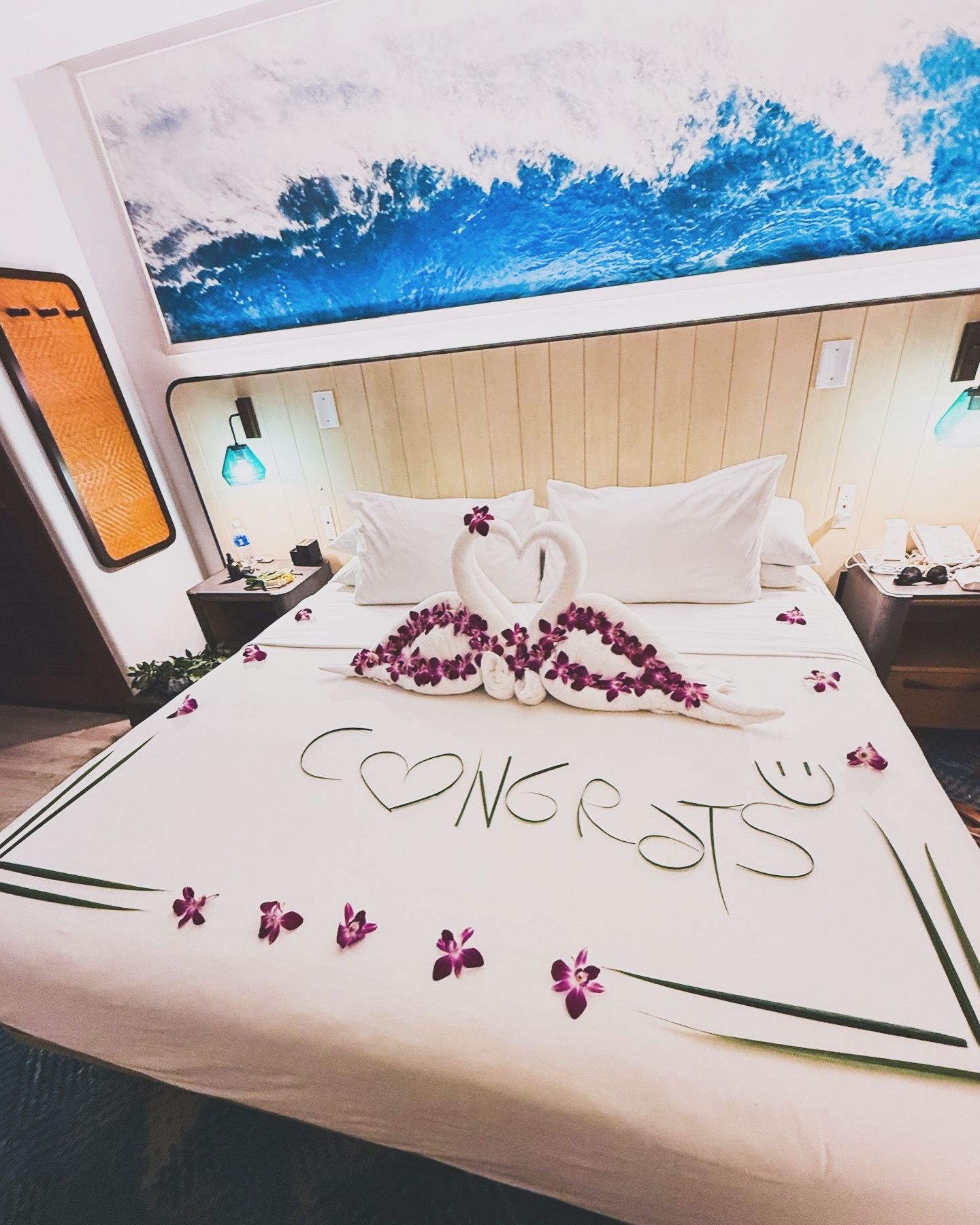 Purple flower petals arranged in a decorative pattern on white hotel bed linens create a romantic atmosphere.