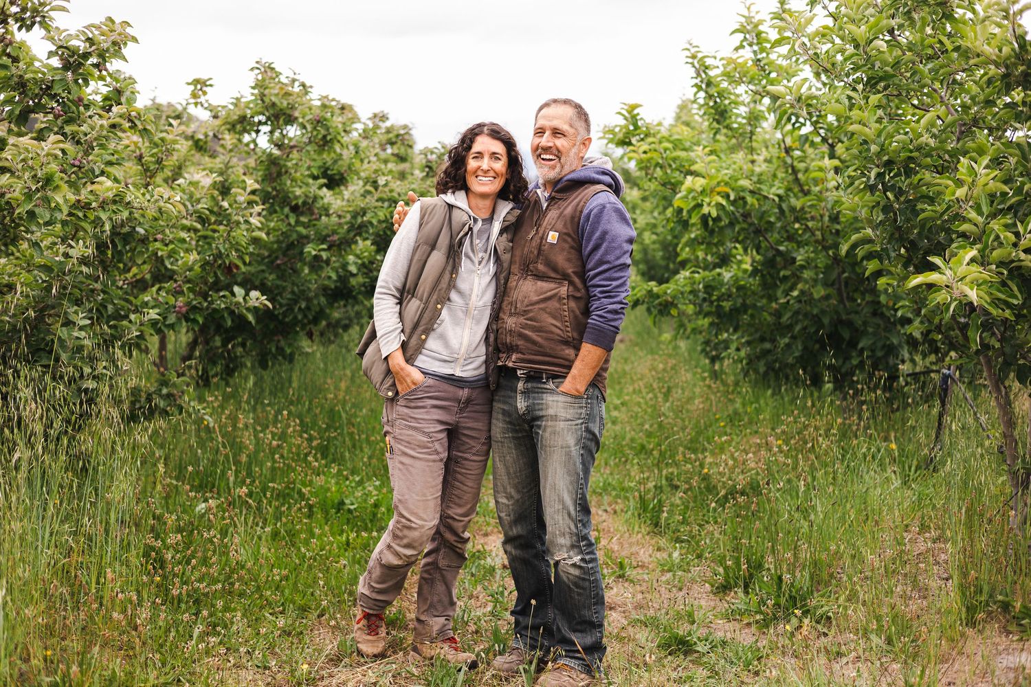 A couple walks together on a dirt path through lush green foliage on a sunny summer day.