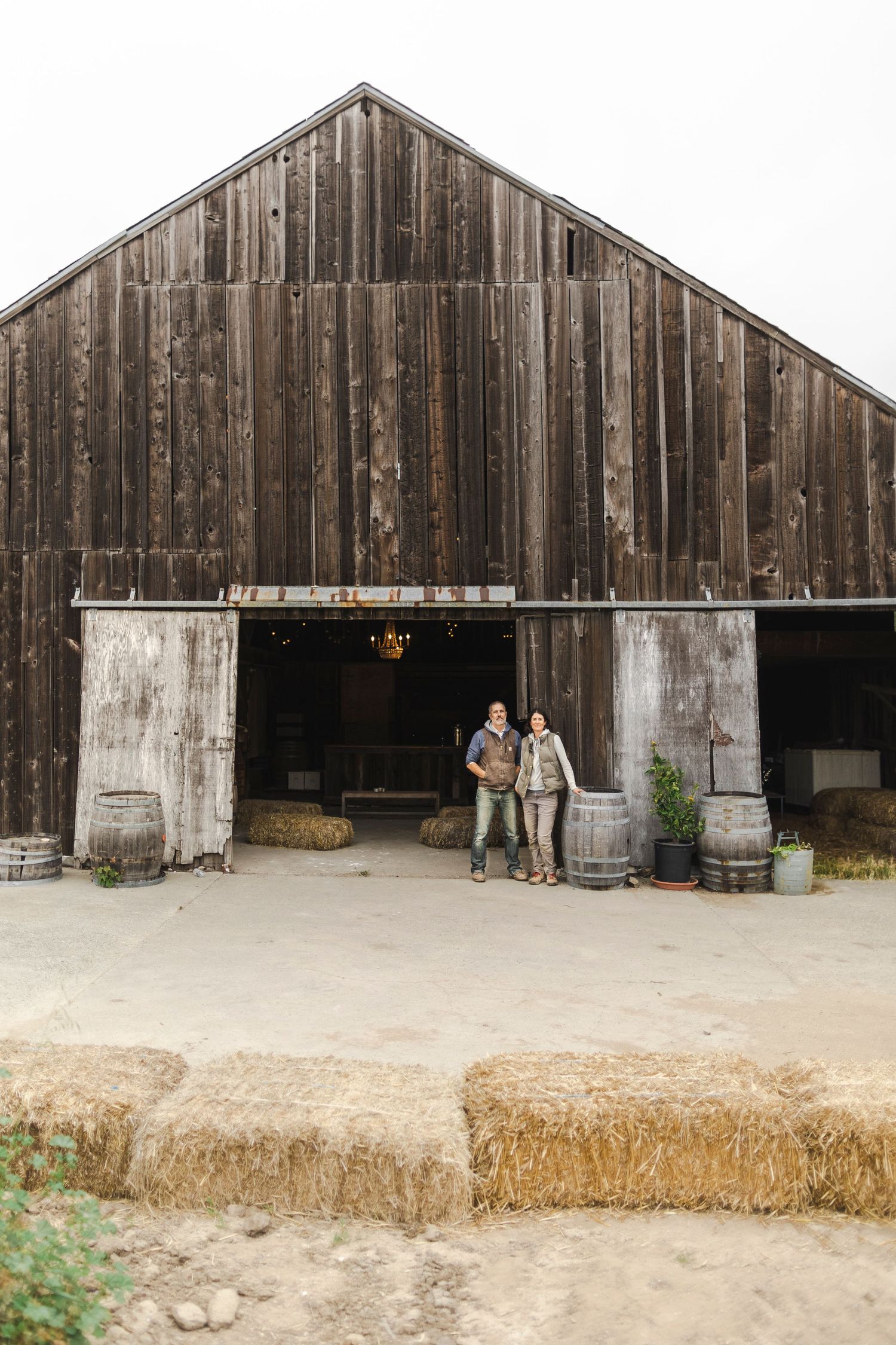 A rustic wooden barn with hay bales and people standing near the entrance in a rural setting.