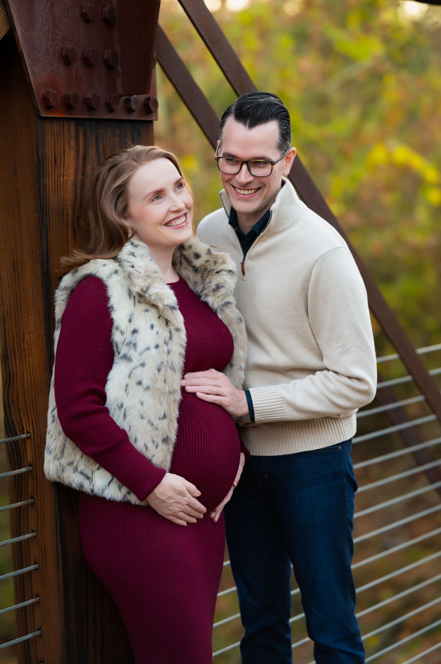 mom-to-be wearing burgundy dress and fur vest leaning on a bridge with father-to-be's hand on bump; parents smiling mid-laughter maternity