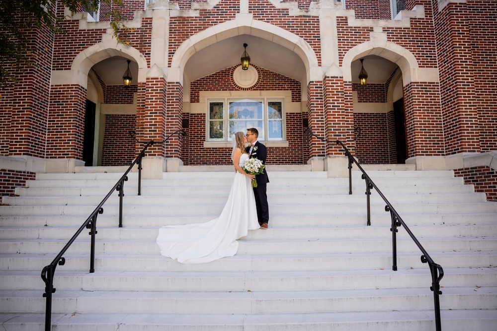 Wedding couple descends stone steps in front of red brick building with arched doorways and white columns.