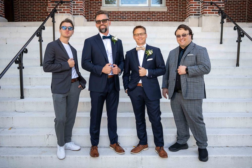 A group of groomsmen in gray suits and brown shoes stand together on white stone steps outside a brick building.