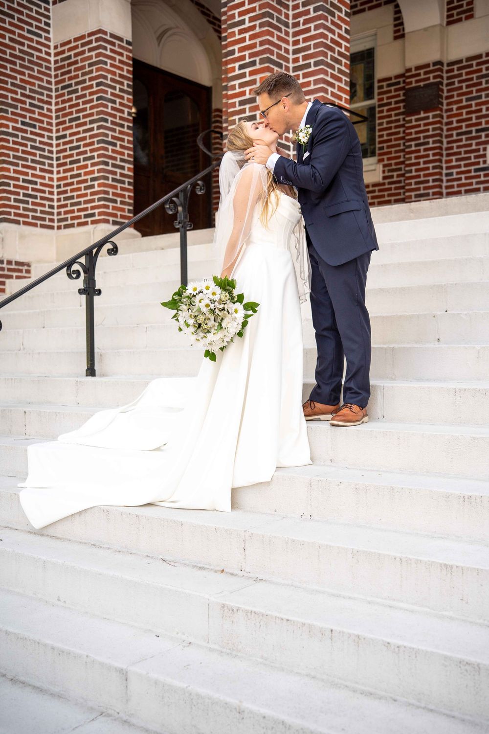 A couple shares a romantic kiss on white stone steps outside a brick church building.