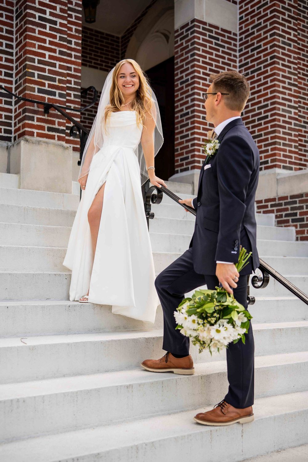 A bride and groom share a joyful moment on church steps with the groom holding a white floral bouquet.