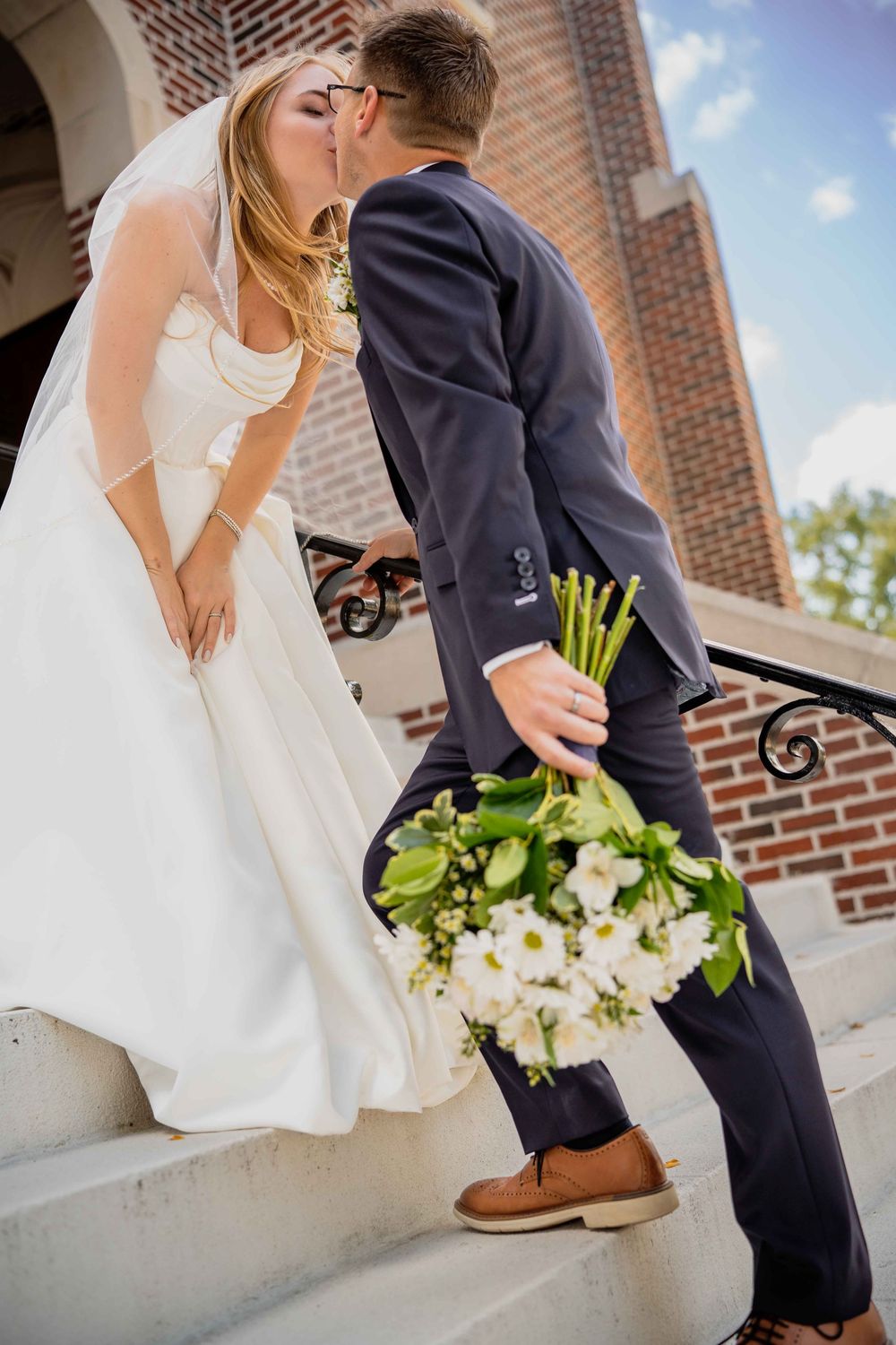 Wedding couple sharing a romantic moment on stone steps with brick building and blue sky in background.