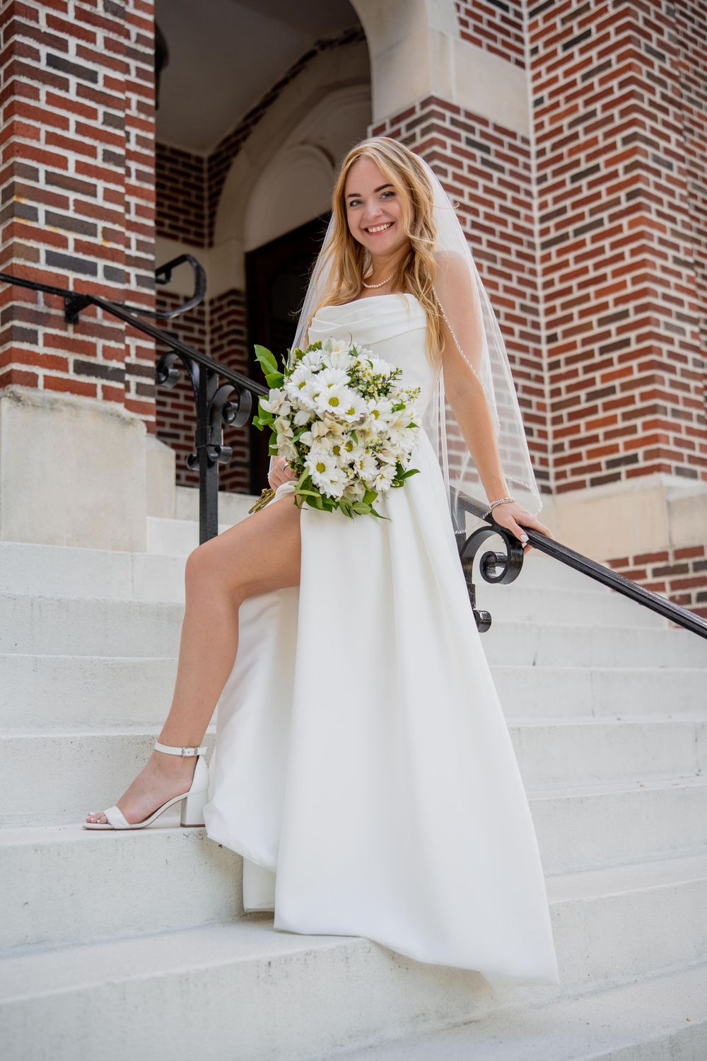 A bride in a white wedding dress sits on stone steps near a brick wall, holding a bouquet of white flowers.