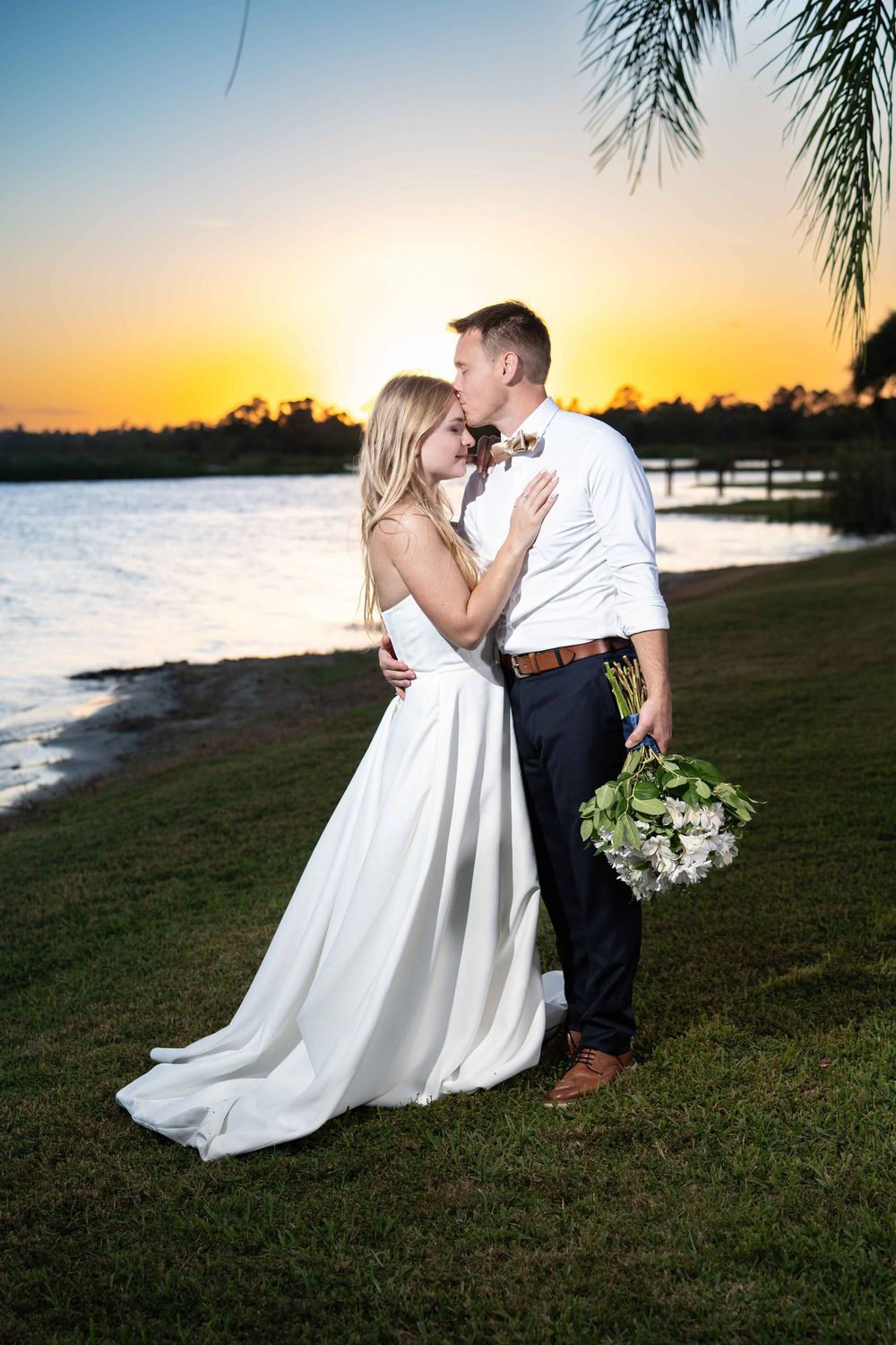 Romantic sunset wedding photos show a couple in elegant attire embracing on grassy waterfront during golden hour.
