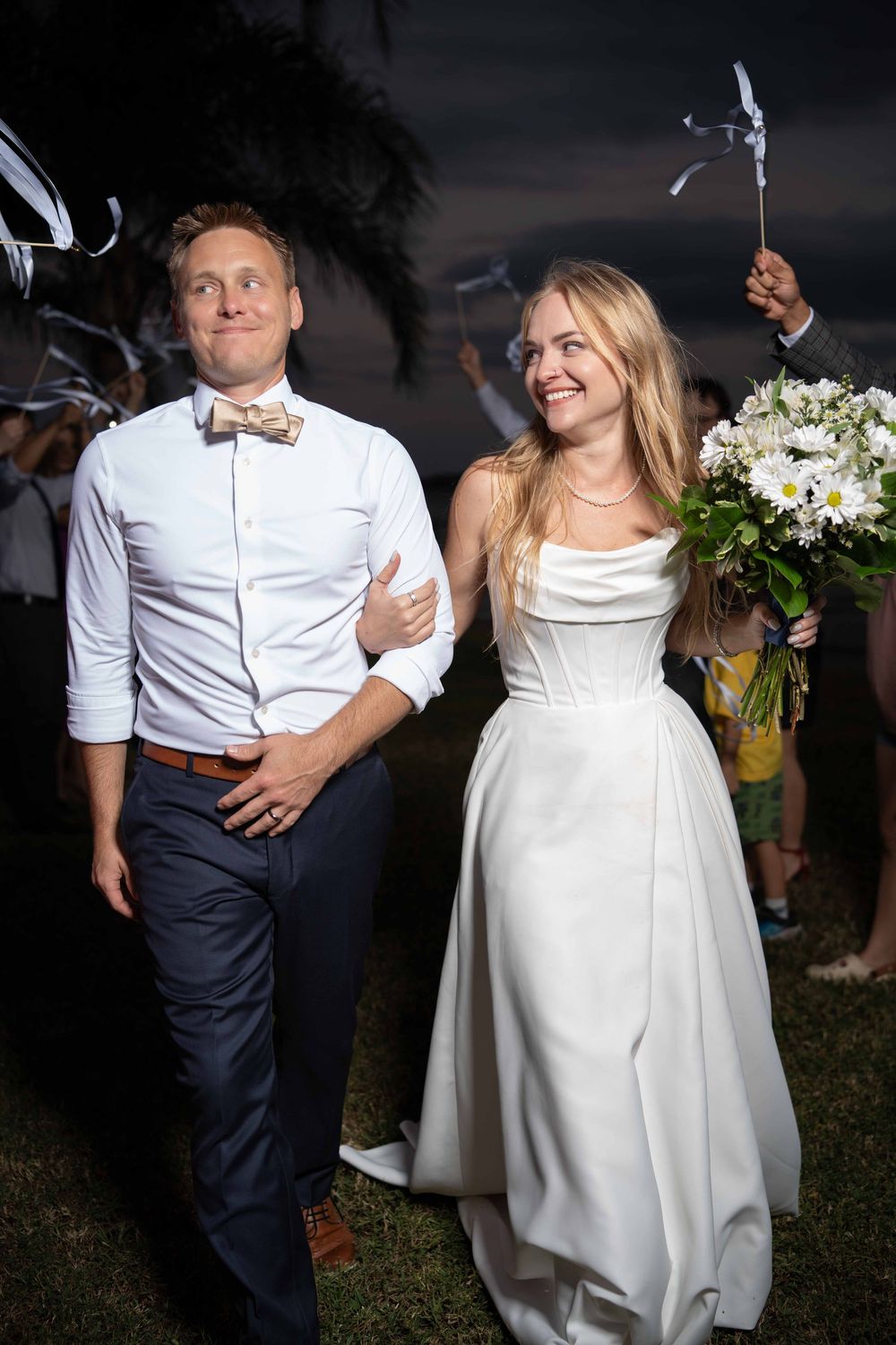 A couple dances together at a wedding reception, with the bride wearing a white dress and groom in navy pants and white shirt.
