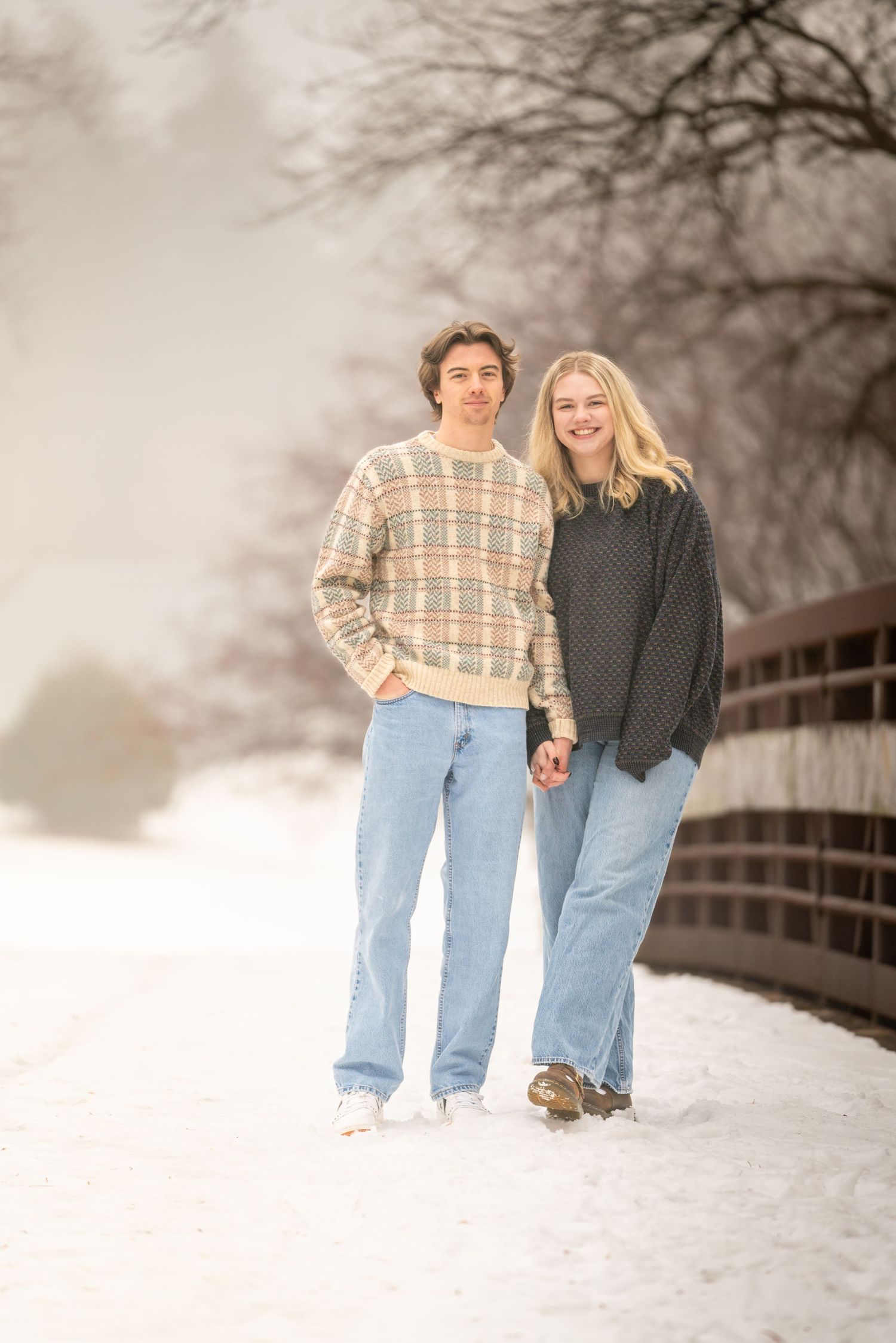 A young couple smiles and holds hands while standing together on a snowy bridge path surrounded by bare winter trees.