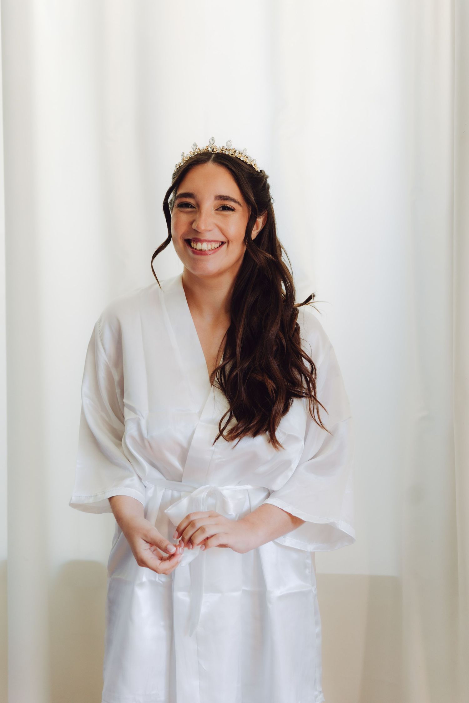Smiling bride wearing gold crown poses in white robe during bridal preparations before wedding.
