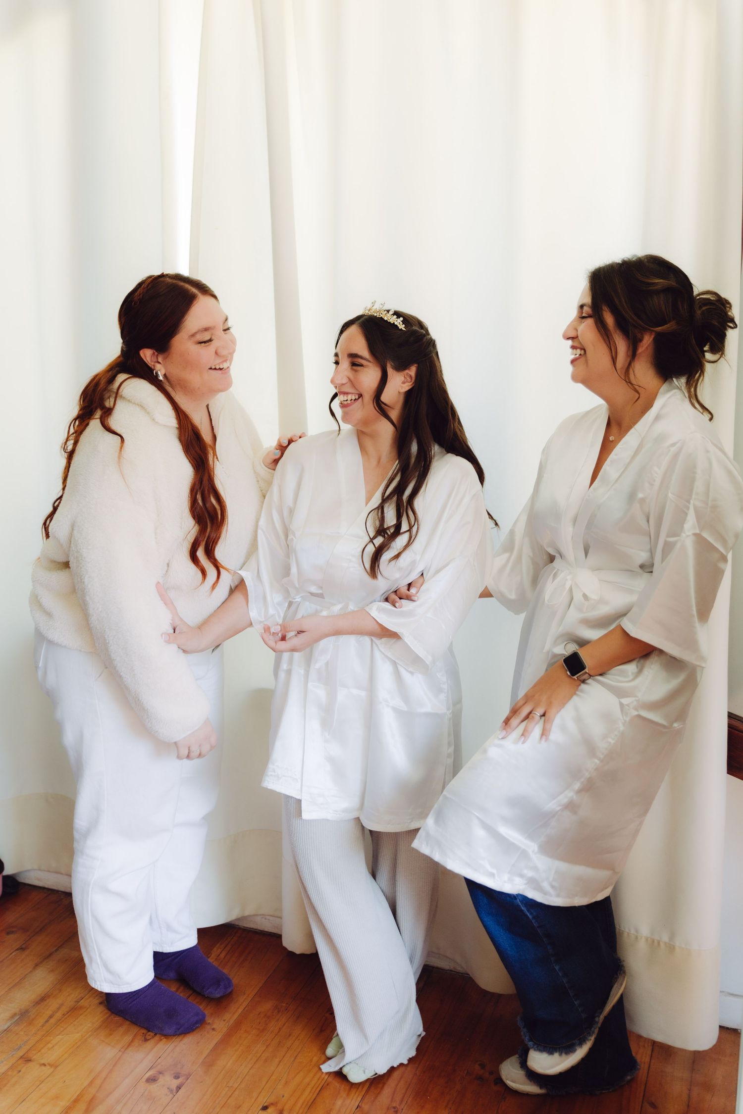Three bridesmaids in white robes laughing together during wedding preparations.